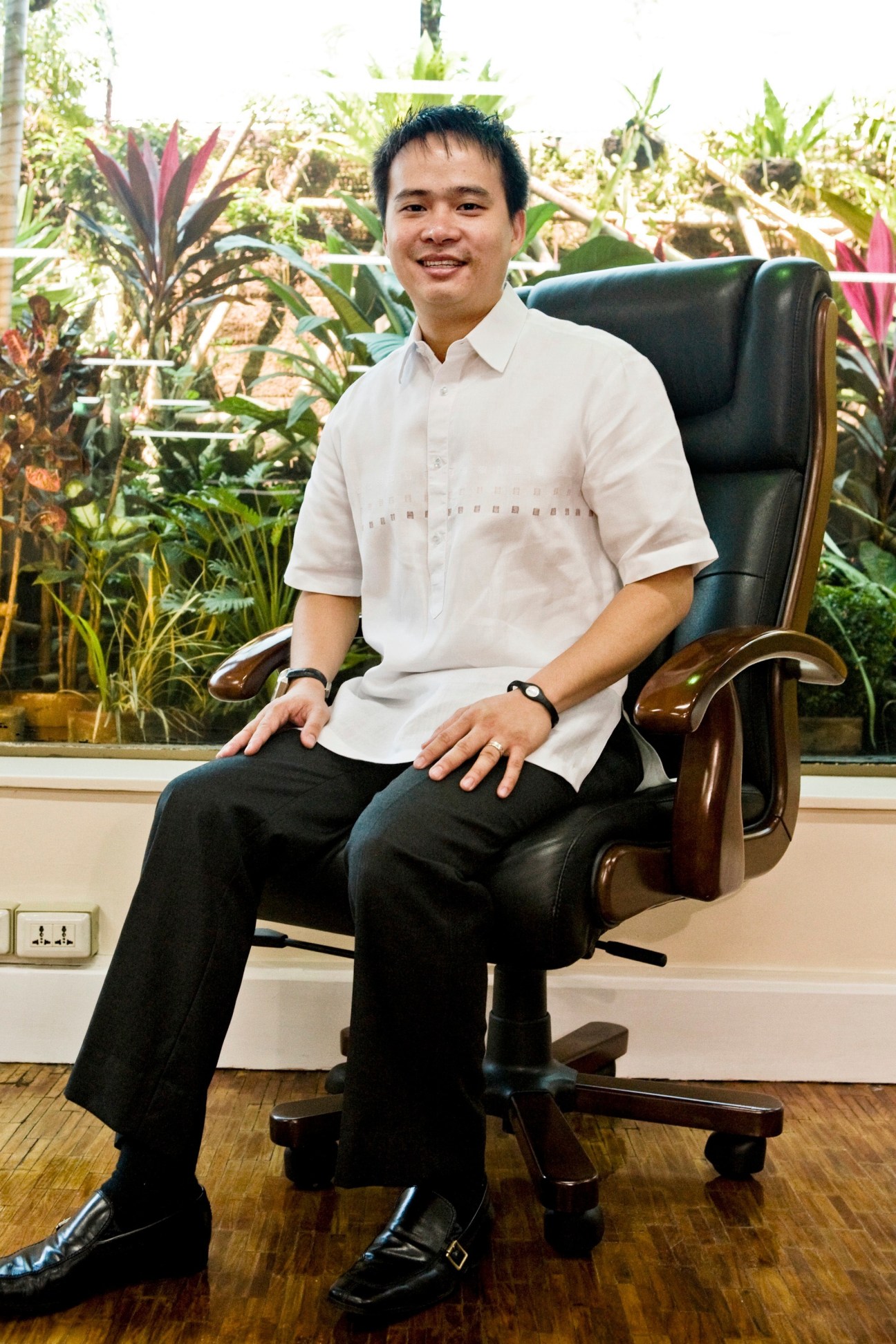 Man in white shirt and black trousers seated in leather office chair with tropical plants behind him