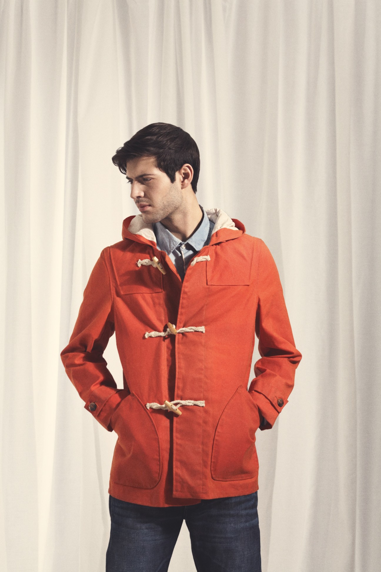 Man wearing a red waxed canvas duffle coat with wooden toggle closures and chambray lining over a plaid shirt.
