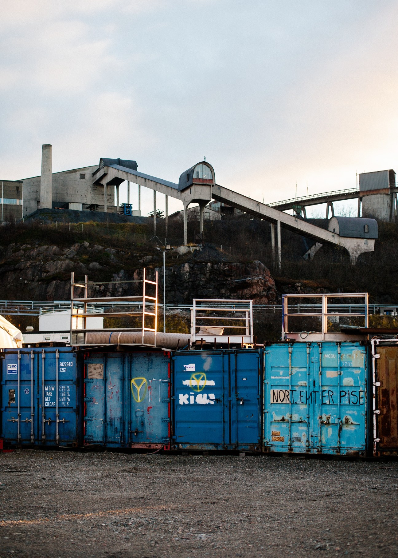 Industrial mining conveyor belt system with blue shipping containers and concrete processing structures in Arctic landscape.