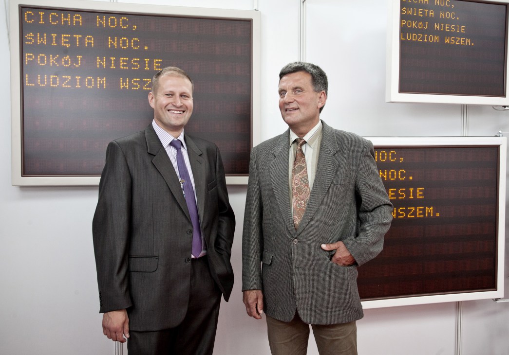 Two men in business suits stand in front of electronic display screens showing Polish text at a trade fair.