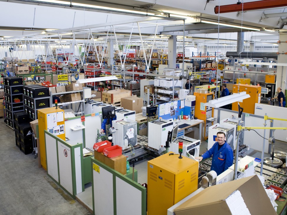 V-Zug factory floor with workers assembling white goods appliances at various workstations.