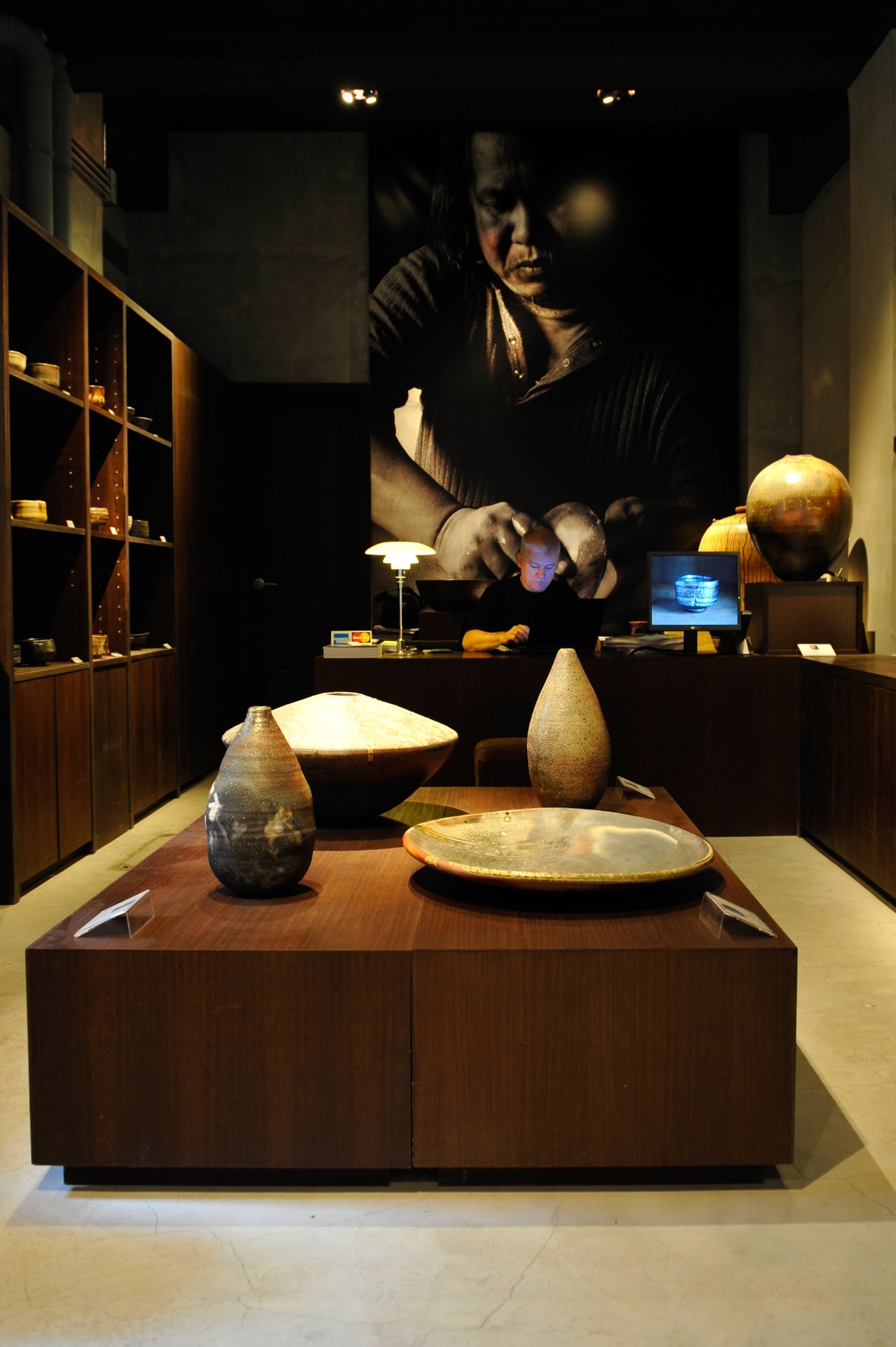 Interior of Earth Home gallery displaying ceramic vessels and pottery on wooden display table with shelving in background.