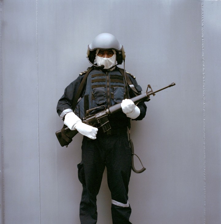 A Singapore Armed Forces recruit in full combat gear and helmet holds a rifle while standing against a plain wall.