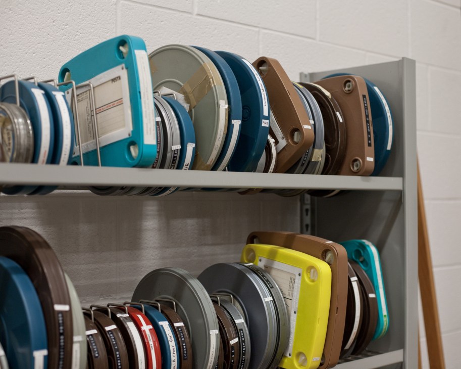 Film reels of Ray and Charles Eames stored on metal shelves in various colors including teal, brown, and yellow.