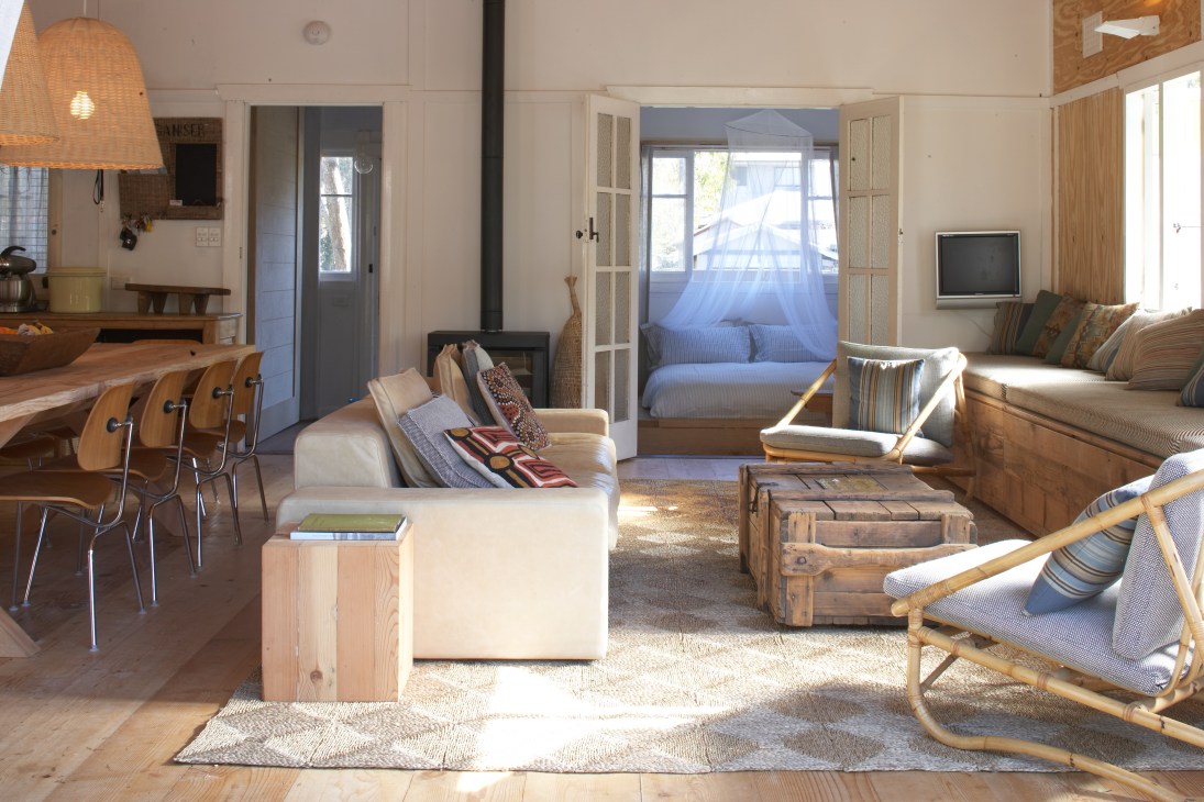 Open-plan beach house interior with wooden furniture, woven pendant light, and natural textiles throughout.