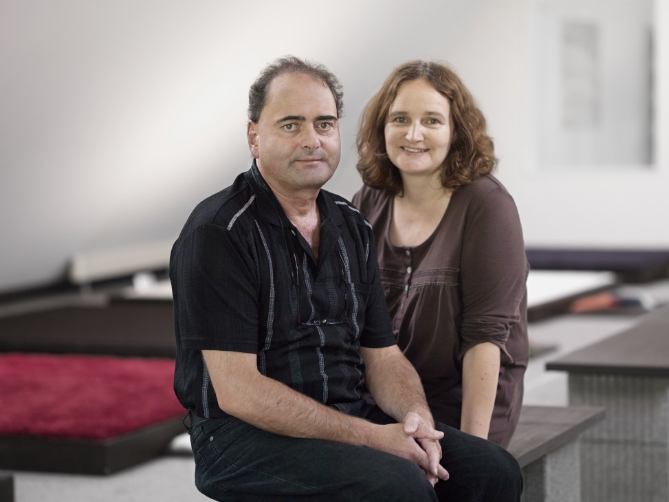 Felix and Doris Kramis pose together in their carpet workshop with finished rugs visible in the background.