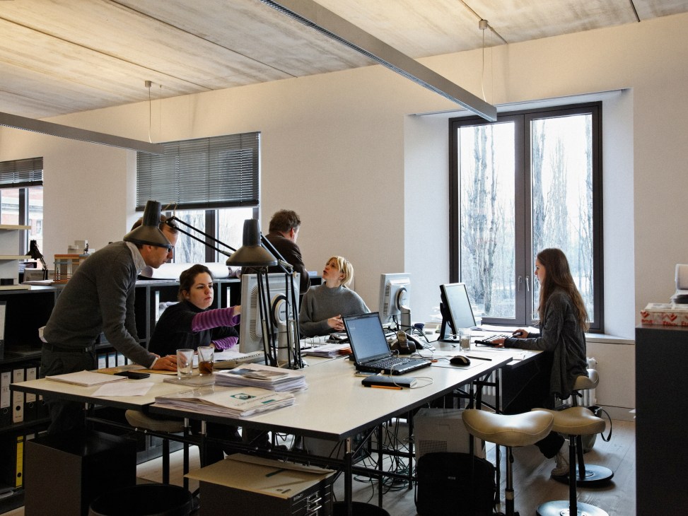 Architects and staff working at desks in a bright studio with exposed concrete ceiling and large windows.