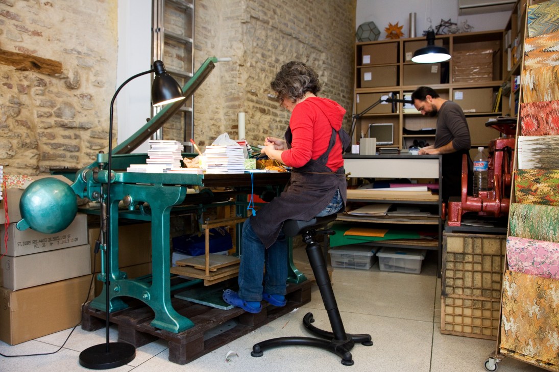 Woman binding notebooks at a large green printing press in Poliedra's workshop.