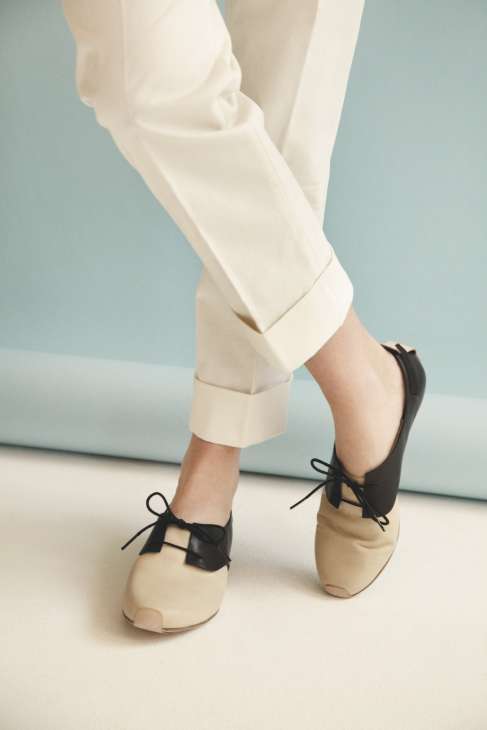 Person wearing cream-coloured trousers and beige Camper shoes with black bows against a blue background.