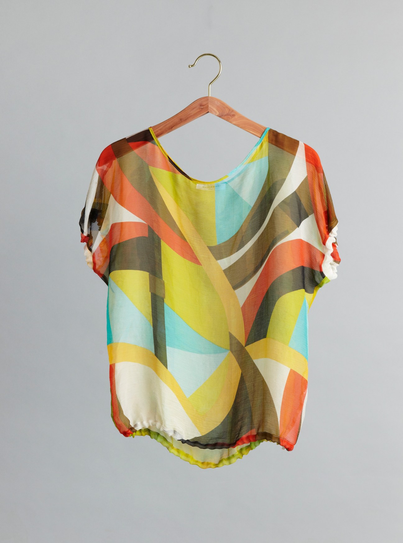 Colorful geometric silk top by Nadia Mari displayed on wooden hanger against gray background.
