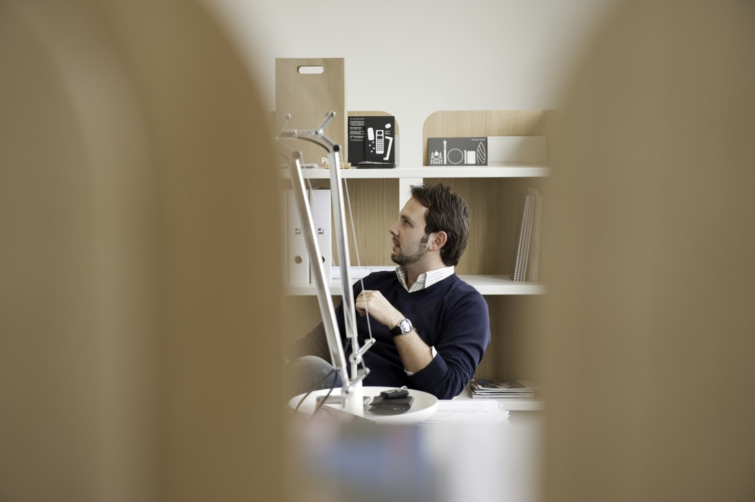Luca Maurer, Punkt's senior product manager, sits at a minimalist desk with a white Tolomeo lamp and shelving displaying the DP 01 phone and other design objects.