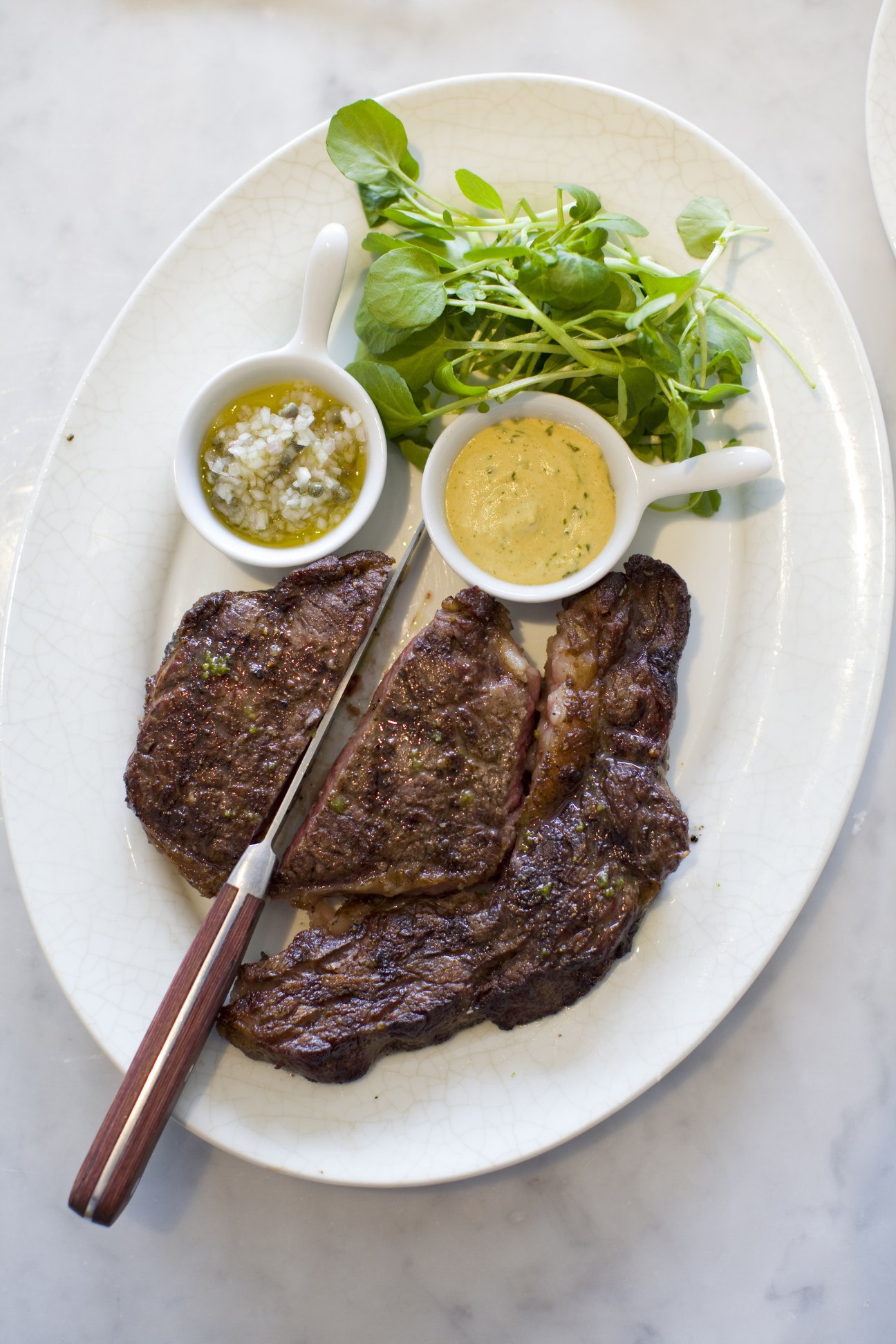 Grilled rib-eye steak with shallot-caper relish, tarragon mustard, and fresh greens on white plate.