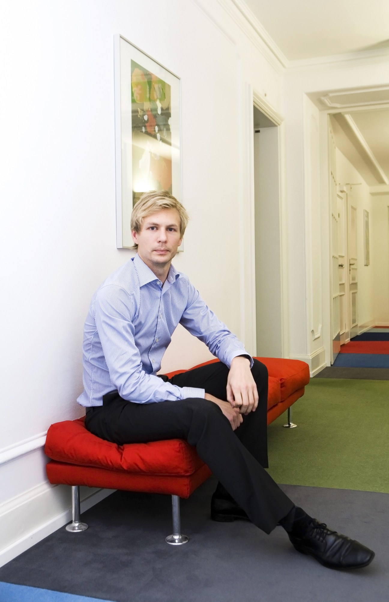 Man in blue shirt sitting on red cushioned bench in modern office corridor