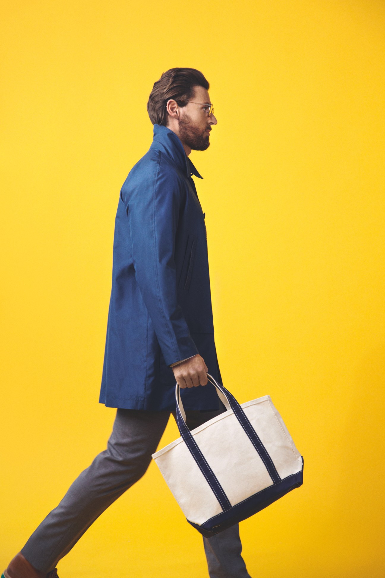 Man in profile wearing a blue Dior Homme coat, Golden Goose shirt, G.T.A trousers, Oliver Peoples glasses, and carrying an L.L.Bean canvas bag against a yellow background.
