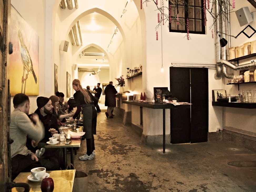 Interior of Snickarbacken 7 concept store café showing white vaulted ceilings, customers seated at tables, and a service counter with shelving.