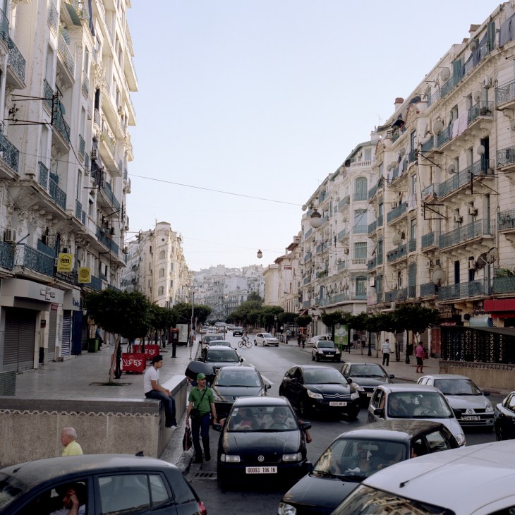 Parisian-style colonial buildings with shutters line a wide street in Algiers with parked cars.