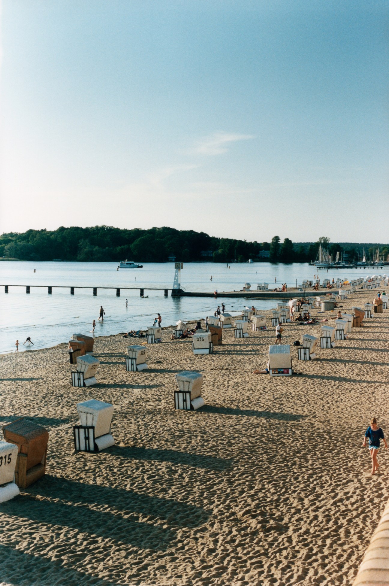 Strandbad Wannsee beach with wooden beach chairs, swimmers, and sailboats on the lake.