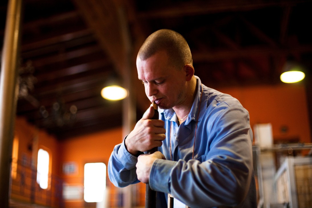 A glassmaker blows through a blowpipe to shape molten glass in a workshop with warm lighting.