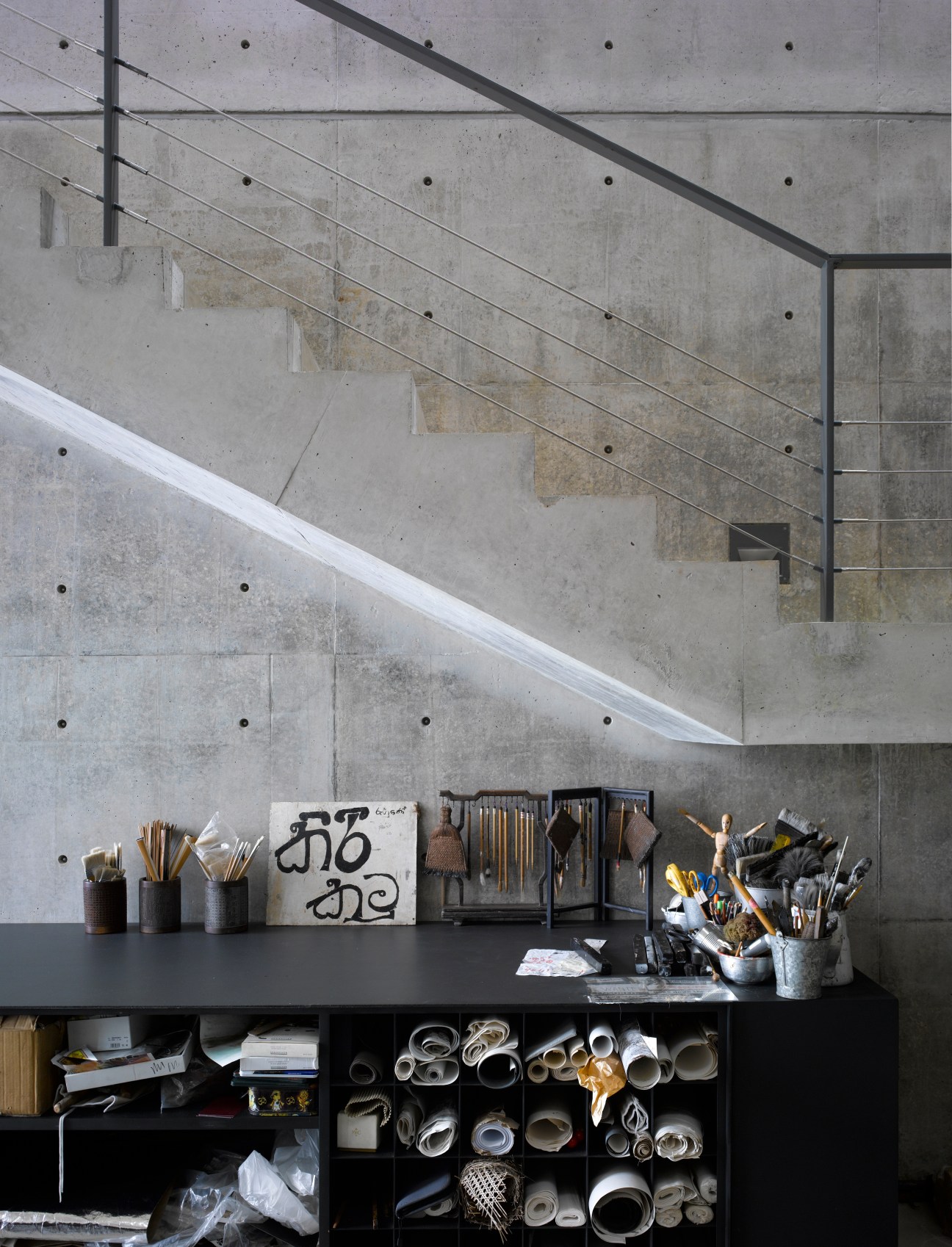 Artist's studio workspace with concrete walls, shelving unit filled with art supplies, brushes in containers, and staircase above.