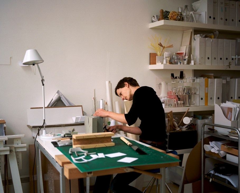 Barbara Etter works at a drafting table with architectural models in a bright studio workspace.