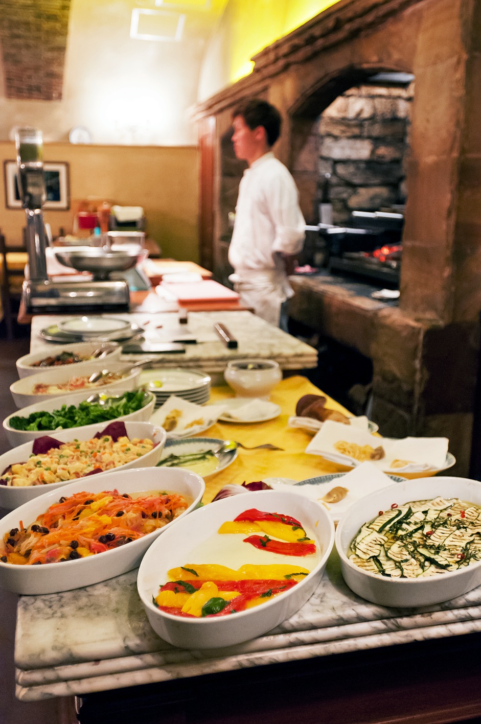 Chef in white shirt works at a traditional Italian restaurant kitchen with various prepared dishes displayed on a marble counter.