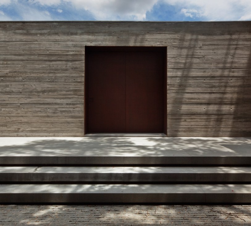 Minimalist entrance of V4 residence with dark wooden door set in weathered wood paneling and concrete platform.