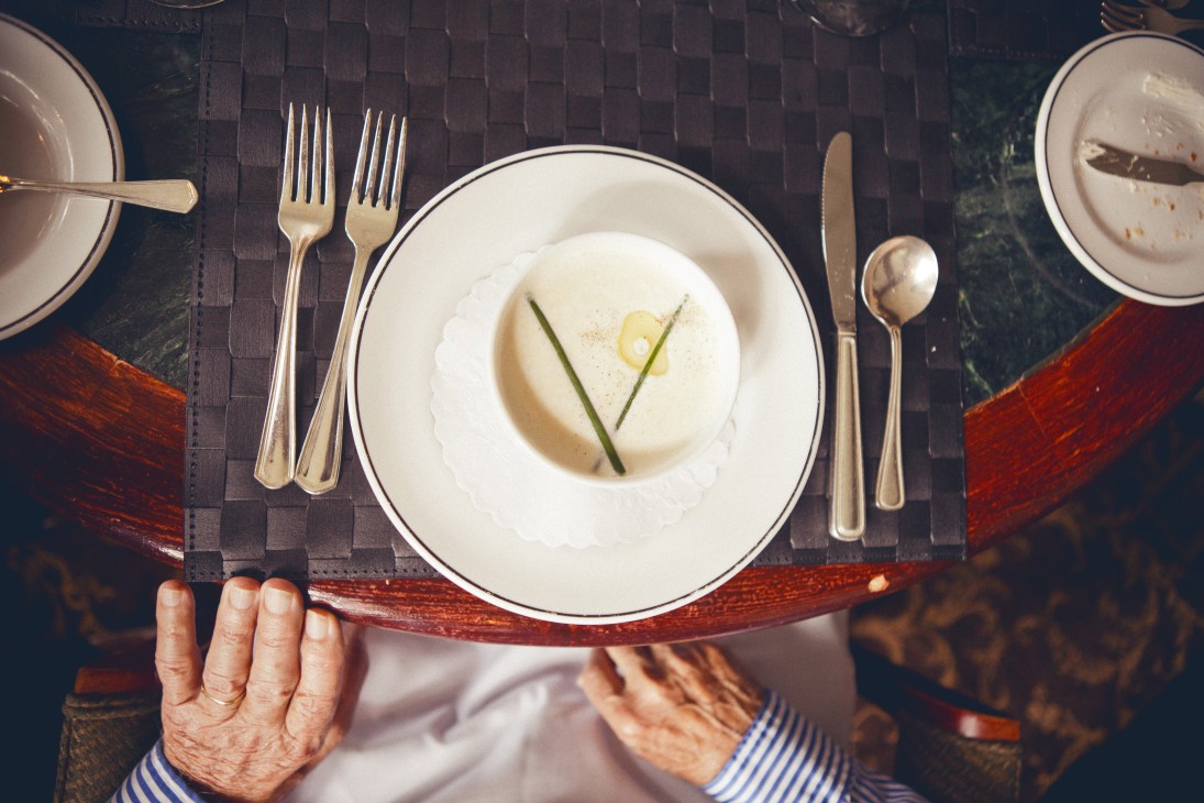 Overhead view of a bowl of chilled vichyssoise soup garnished with chives, served on a dark textured placemat with silver cutlery.