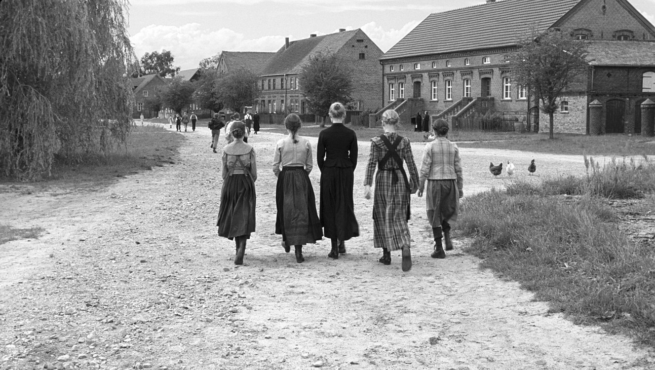 Black and white film still showing five children walking away from camera toward rural German village buildings.
