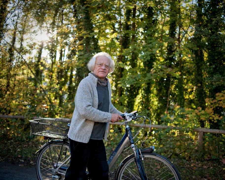 Jürgen Thiele, an elderly man with white hair and glasses, stands with his blue Koga electric bike in a wooded area.