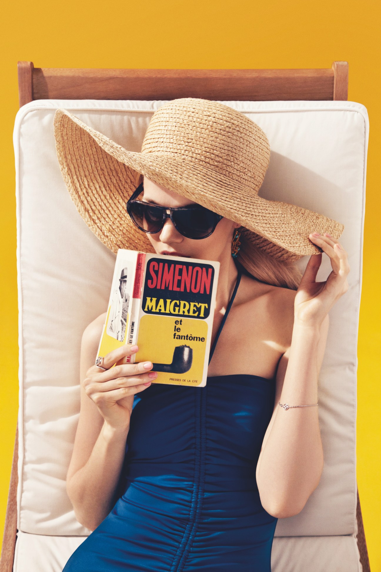 Model wearing Chanel sunglasses and Ralph Lauren straw hat on lounger holding a Simenon book.