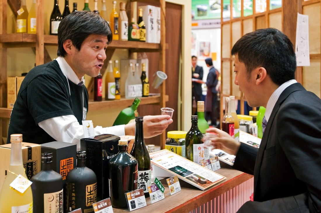 Two men in business suits sample locally made alcoholic drinks at a wooden bar with bottles displayed behind them.