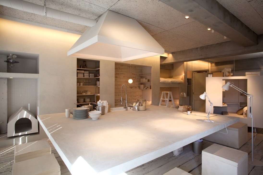 Modern minimalist kitchen and living space with white central island, wooden shelving, concrete ceiling, and integrated appliances.