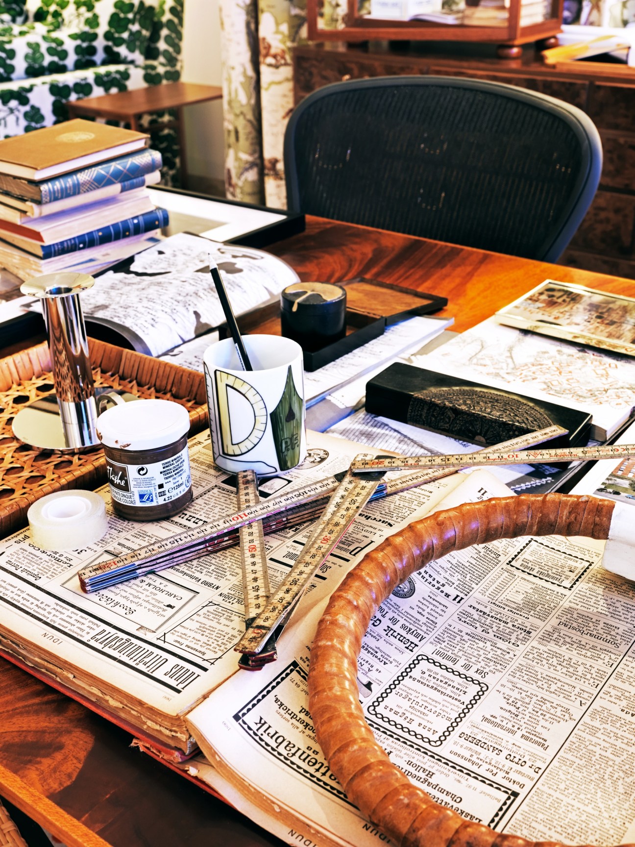 A wooden desk cluttered with design materials, books, patterned fabric samples, and sketching tools.