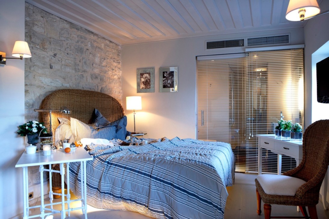 A bright, modern hotel bedroom with wicker headboard, blue striped bedding, stone accent wall, and glass doors opening to a terrace.