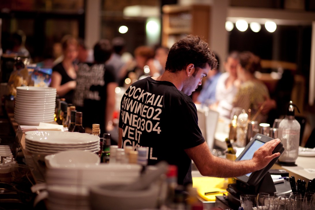 Bartender in black t-shirt preparing cocktails at a busy restaurant bar counter with white plates and bottles.