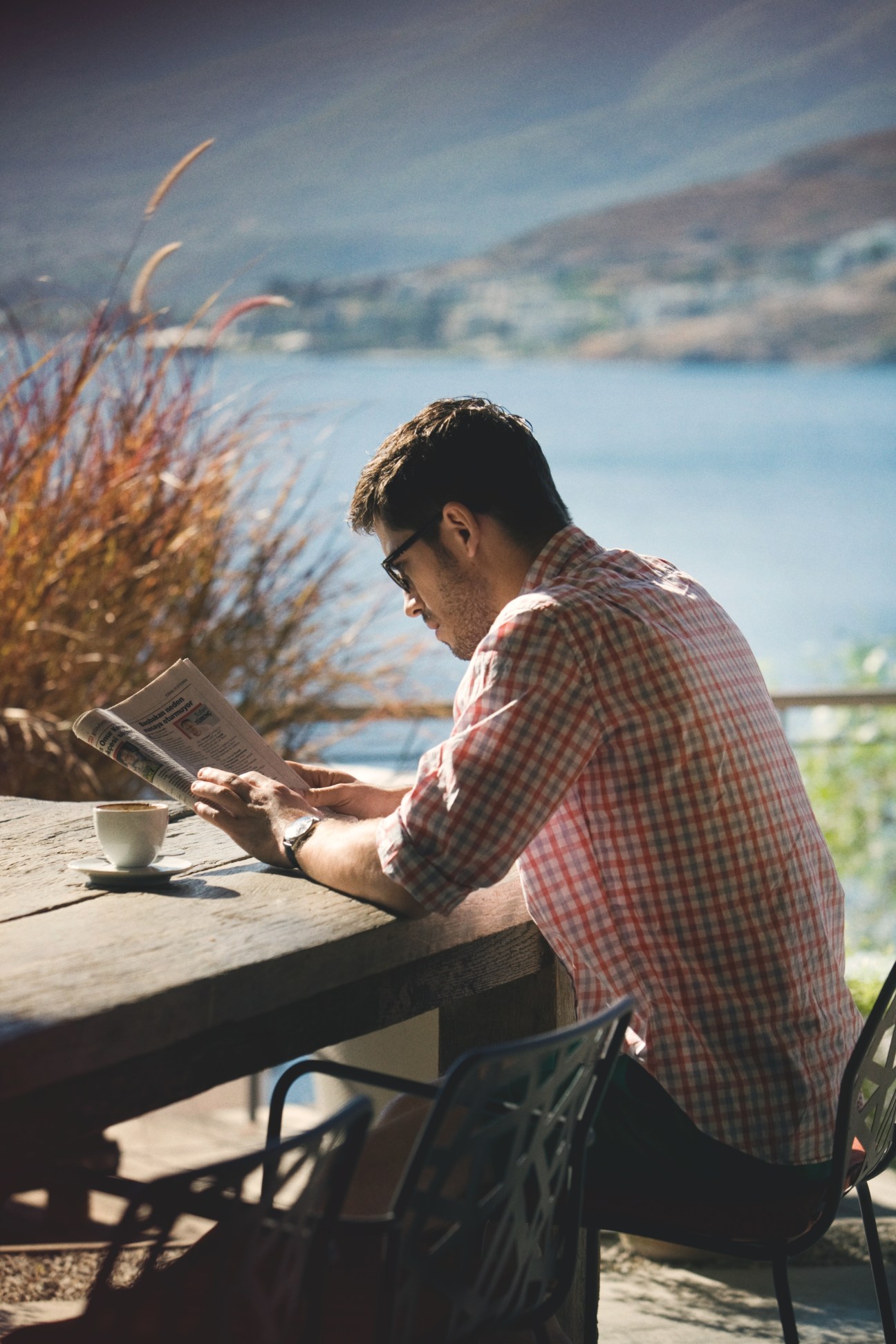 Man in Persol sunglasses and J.Crew checkered shirt reading newspaper by coastal overlook in Bodrum.