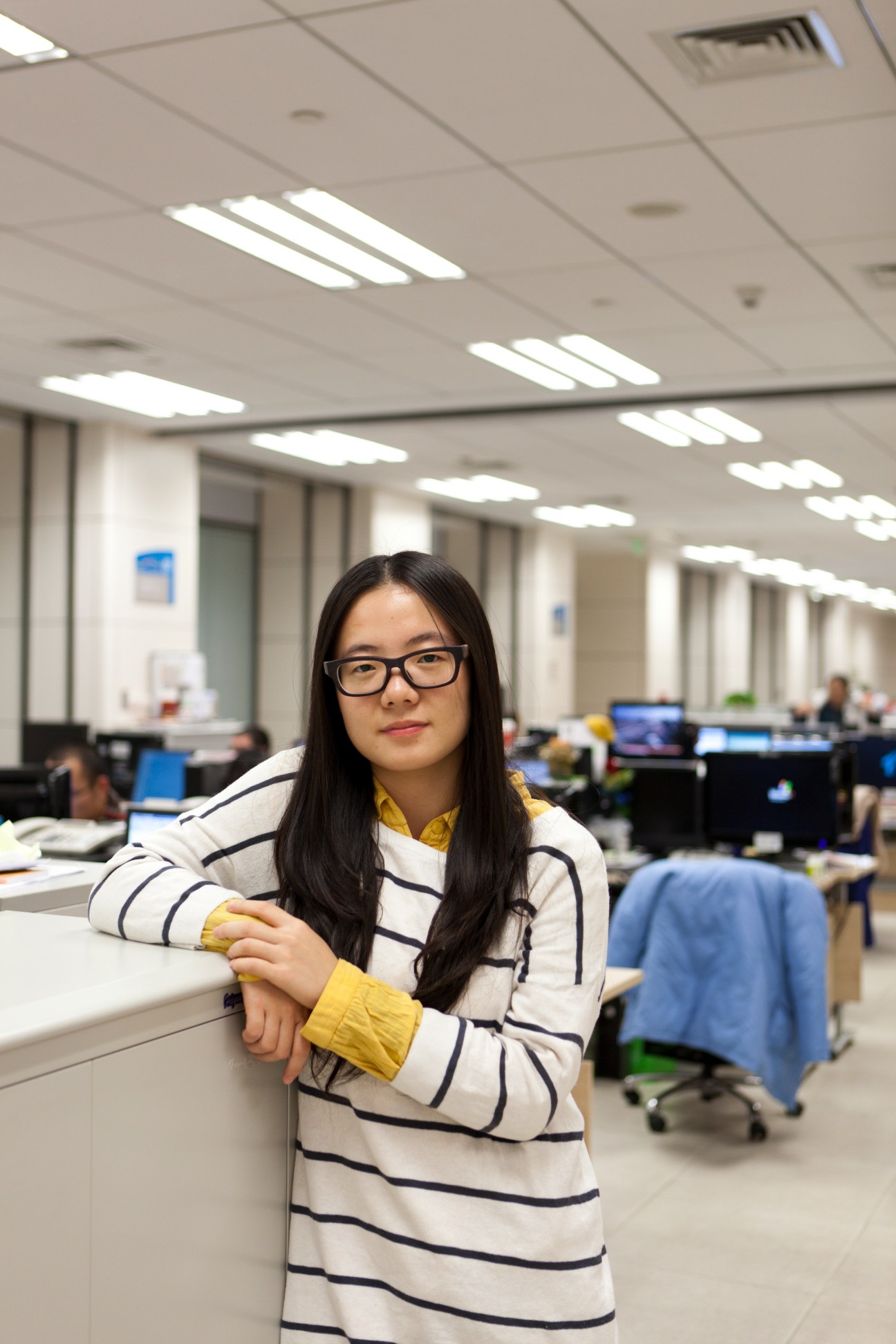 Du Yinghan, animation assistant director, poses in a modern office newsroom with fluorescent ceiling lights and workstations visible in the background.
