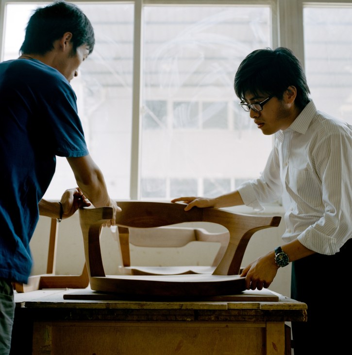 Two men examine wooden furniture components at a workbench near a bright window.