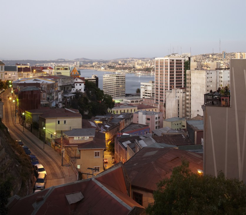 Evening view of Valparaíso's hillside cityscape with bay waters, mixed architecture, and Hotel Fauna terrace visible on right.
