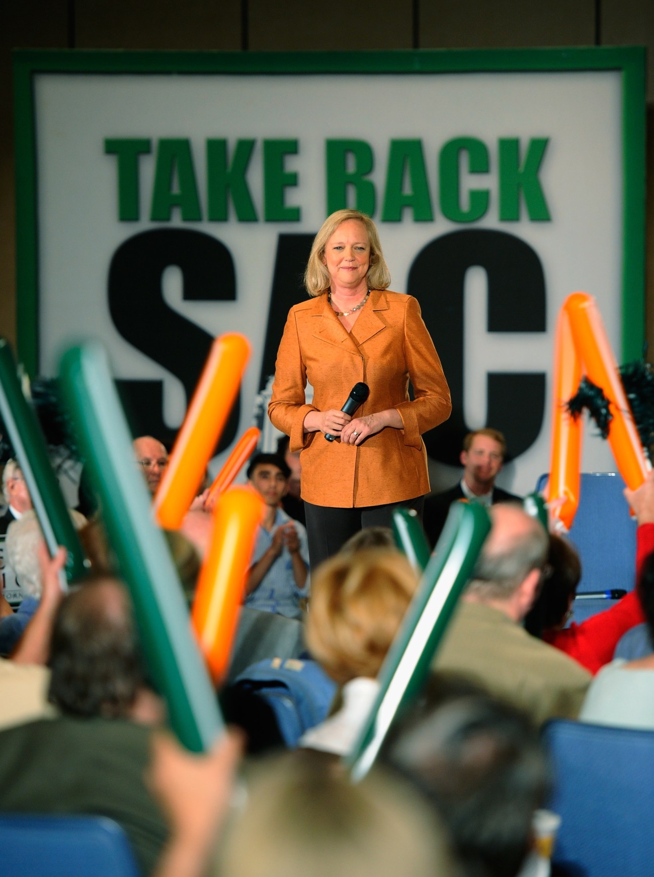 Woman in orange blazer speaking at podium with "Take Back" sign behind her during political rally.