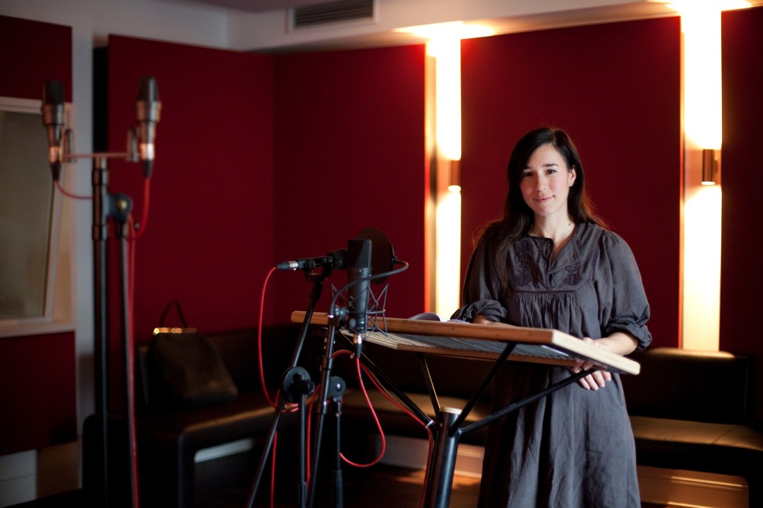 Anke Kortemeier stands in a professional recording studio with red soundproof walls, holding a script beside a microphone and stand.