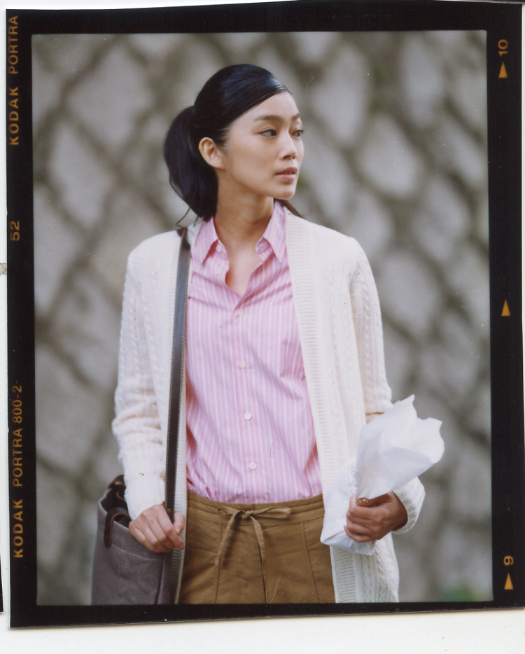 Woman wearing a cream cardigan, pink striped shirt, and olive trousers standing outdoors.