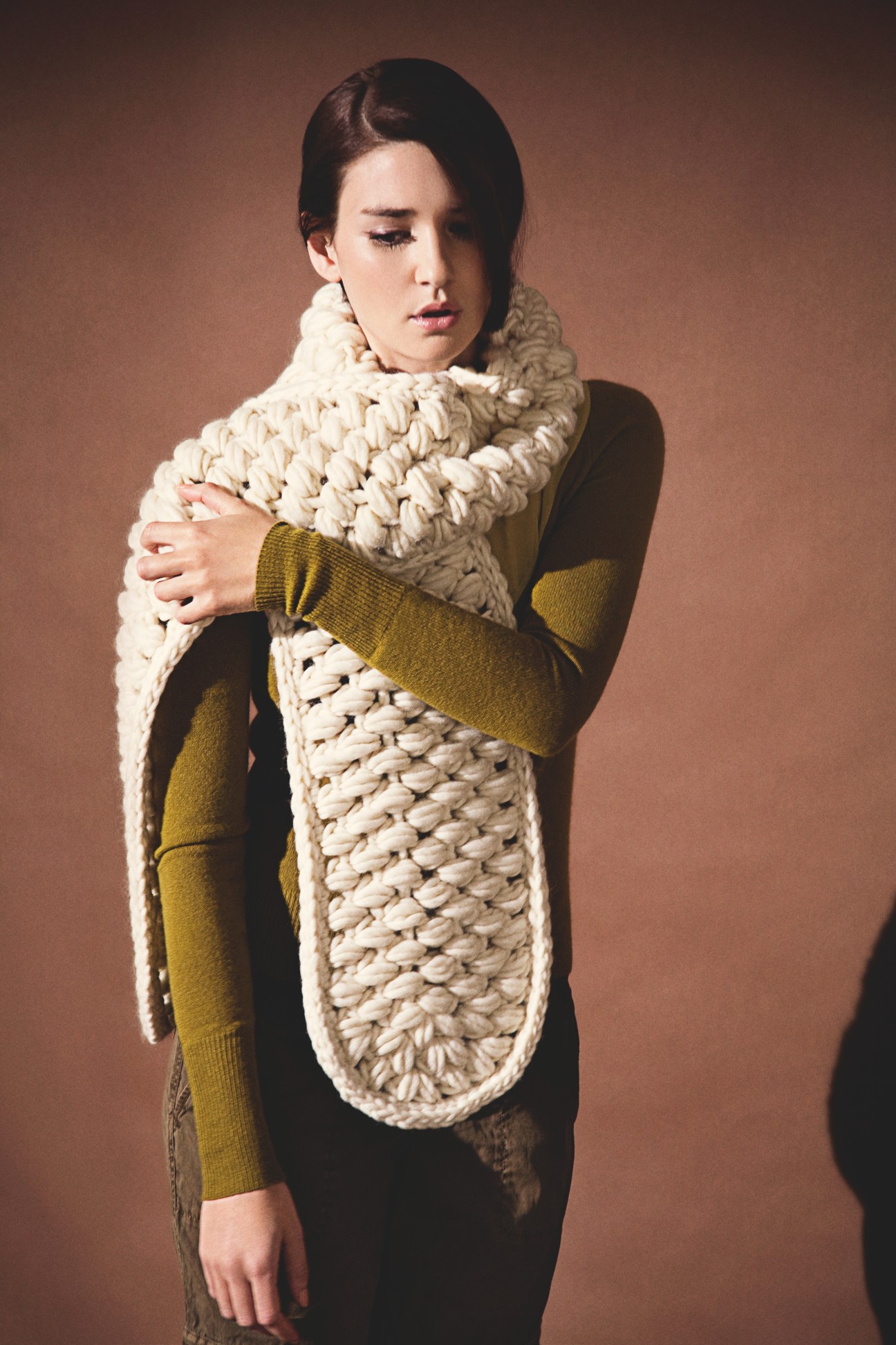 Woman wearing a cream chunky knit scarf with a mustard yellow sweater against a brown background.