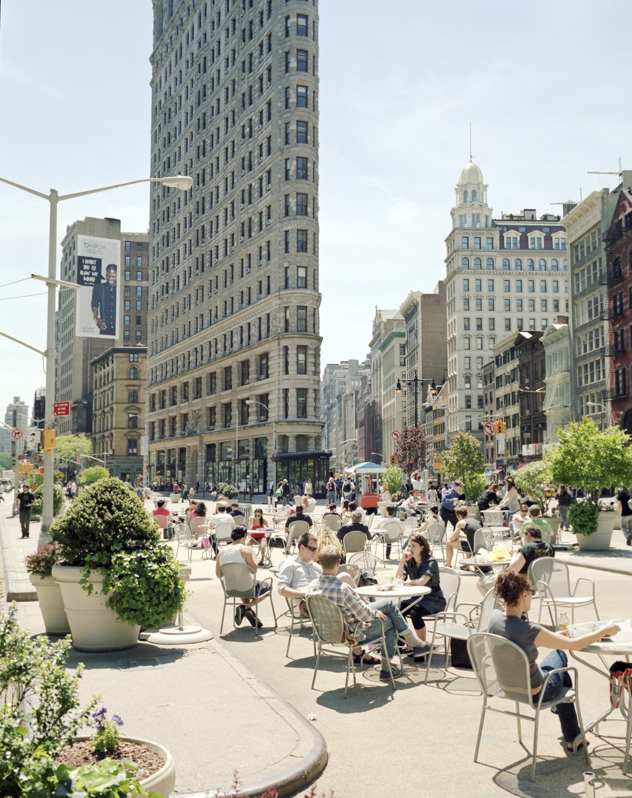 People seated at movable chairs and tables in a sunny New York City plaza surrounded by tall buildings.