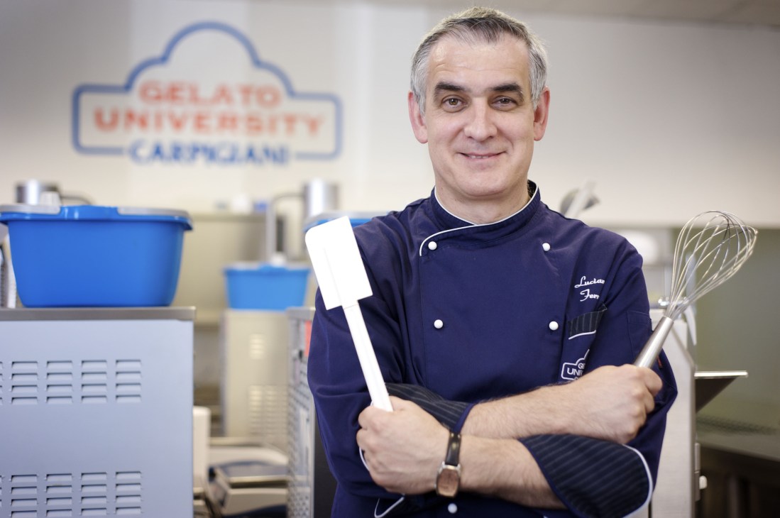 Gelato maestro Luciano Ferrari in chef's jacket holding serving tools in Carpigiani Gelato University kitchen.