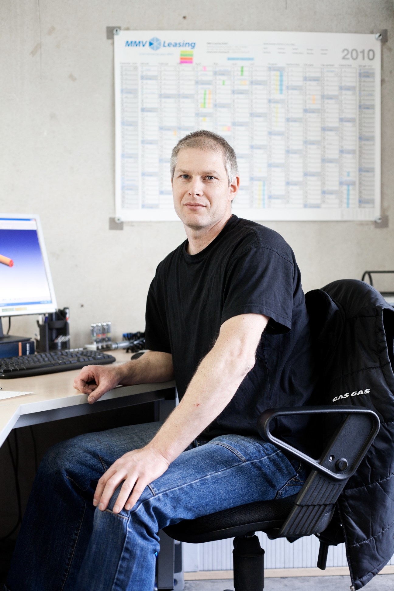 Man sitting at desk with computer and calendar on wall behind him.