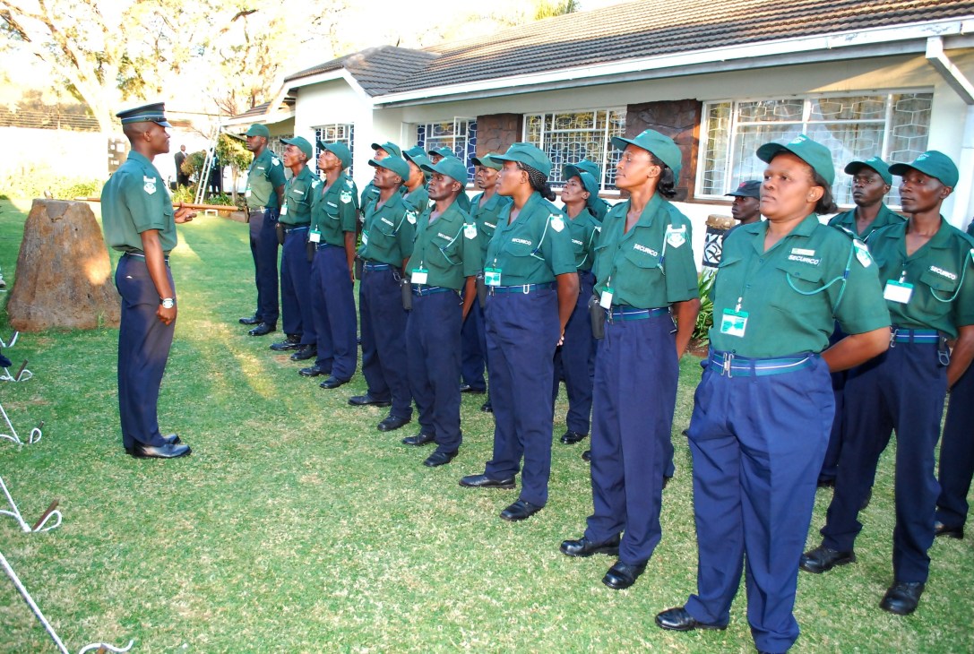 Female security guards in green uniforms and teal caps stand in formation on a lawn during a briefing.