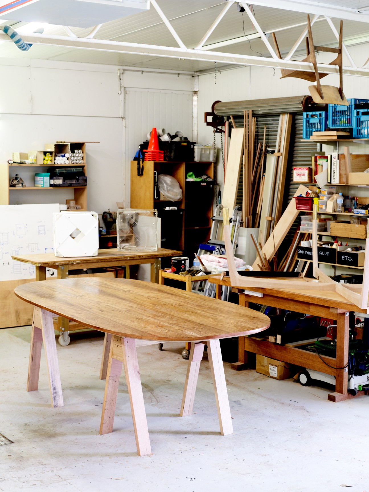 Wooden table with oval top and pale pink legs in a bright workshop filled with timber and tools.