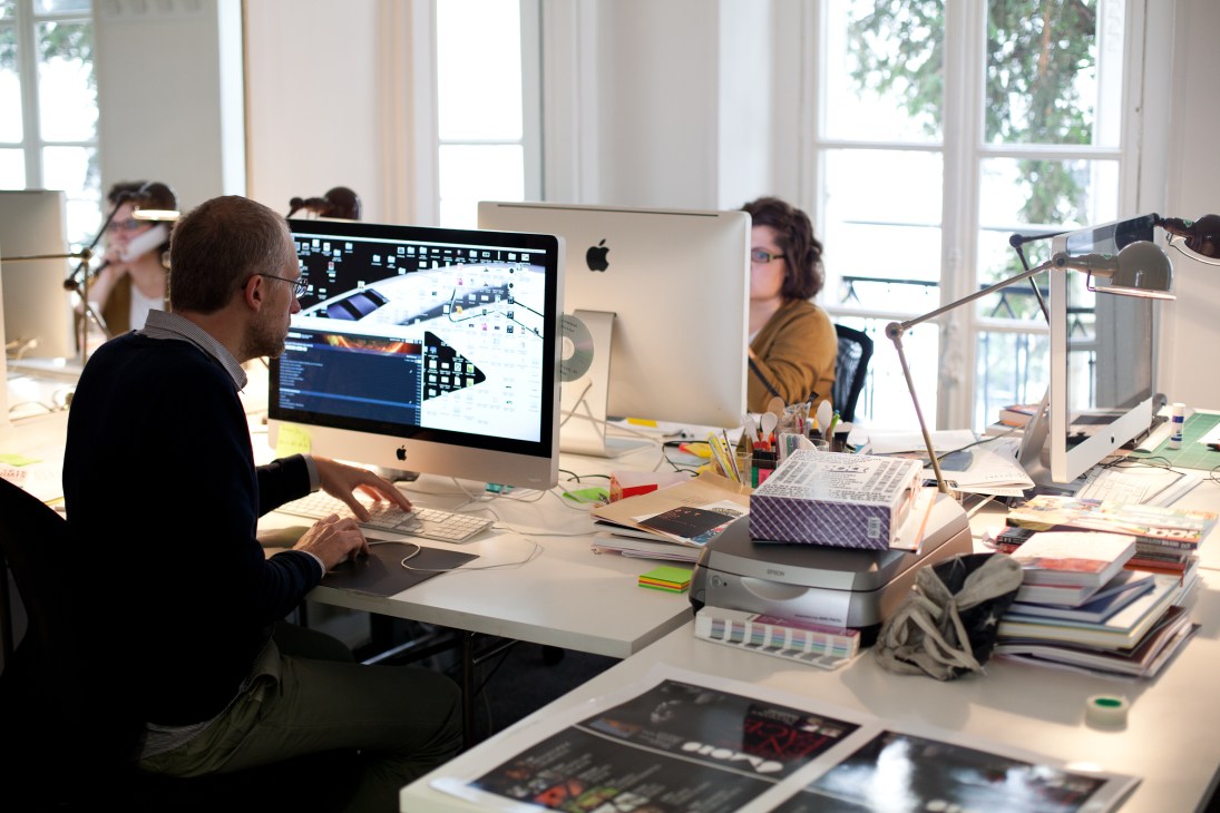 Quintin Leeds working at a desk in a bright art department with large windows, using an iMac computer.