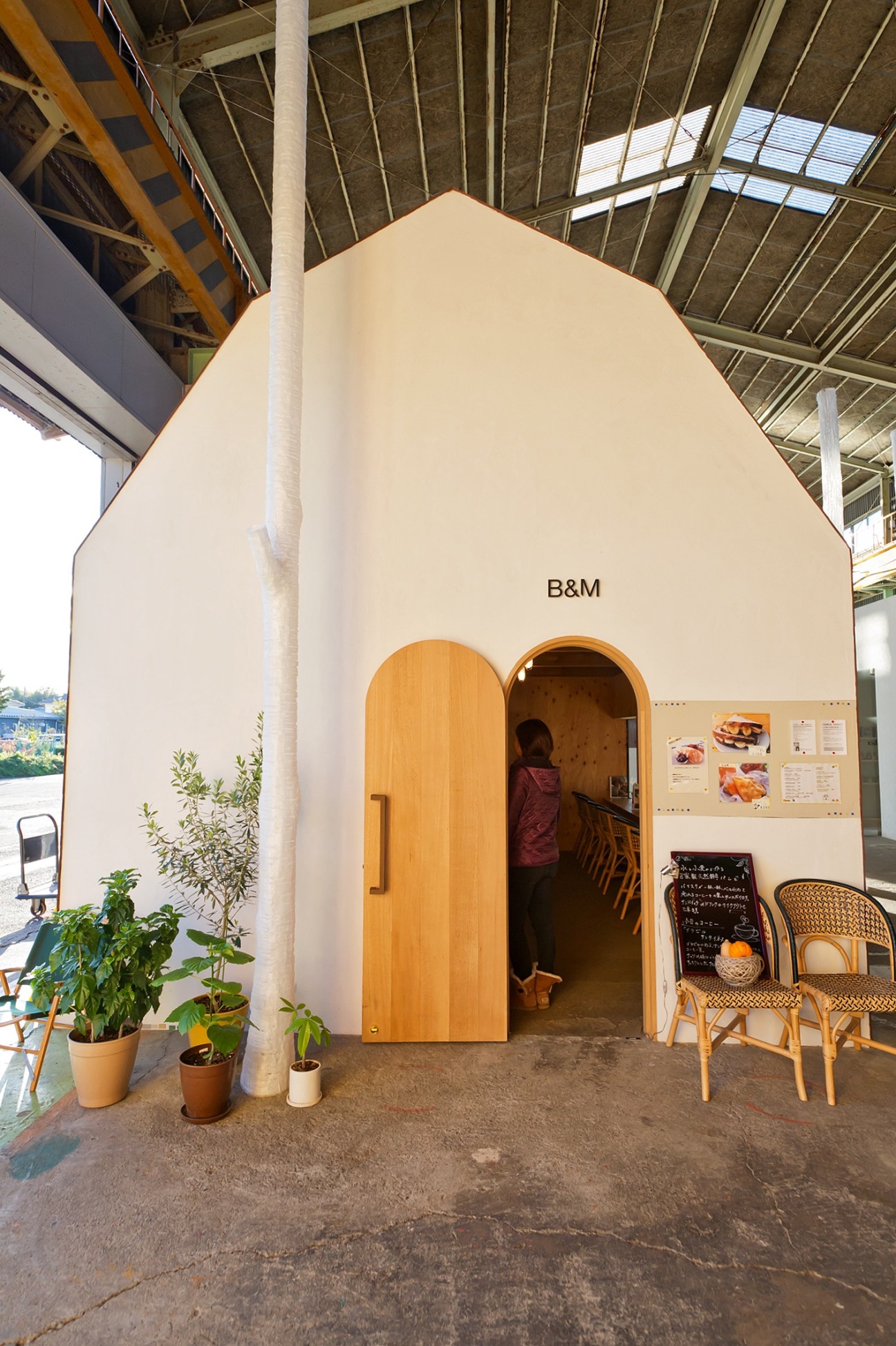 A white building with arched wooden door labeled B&M, potted plants outside, and chairs visible to the right, set in an industrial warehouse space.